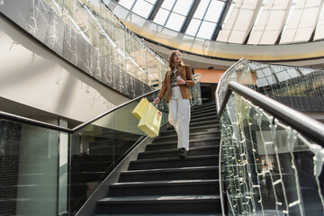 Cheerful woman with cellphone holding shopping bags on stairs in mall.