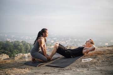 Fototapeta na wymiar Strong caucasian man doing abdominal crunches on yoga mat while african woman holding his bare feet. Multiracial active couple spending free time for outdoors activity.