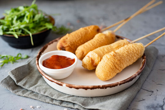 Quick snack: corn dog on a stick. Sausage in batter on a long skewer on a white plate. Close-up
