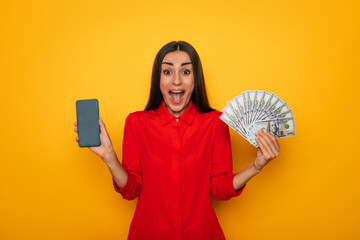 Attractive excited smiling brunette holds bunch of money and smart phone in hands while posing on yellow background