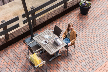 Overhead view of stylish woman sitting near gadgets, dessert and shopping bags on terrace.