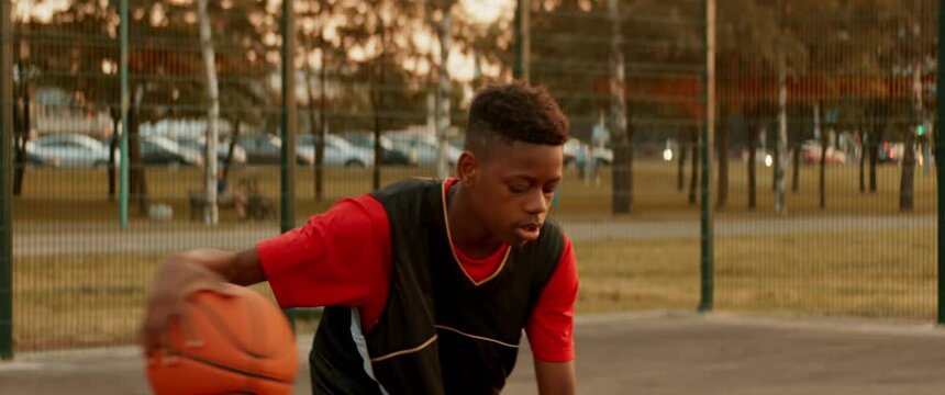Black African American teenager kid boy playing basketball alone on an outdoor court in the evening. High quality 4k footage