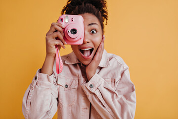 Studio shot of amazed girl holding camera. Front view of african american woman taking photos on yellow background.