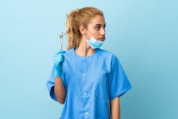 Young woman dentist holding tools over isolated blue background looking side