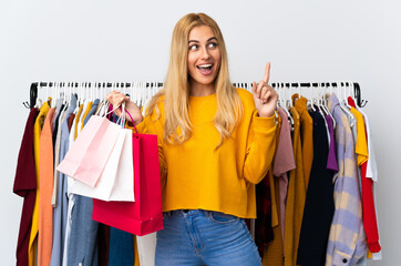 Young Uruguayan blonde woman in a clothing store and holding shopping bags intending to realizes the solution while lifting a finger up