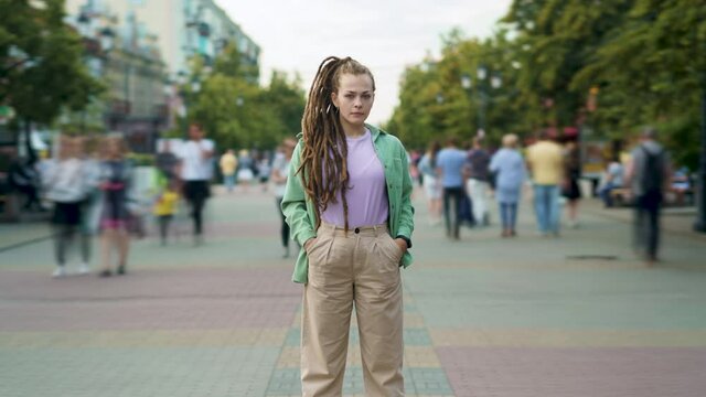 Zoom-in time lapse of unique girl with dreadlocks hairdo standing outdoors in city street among crowd pf people rushing around. Youth and life concept.
