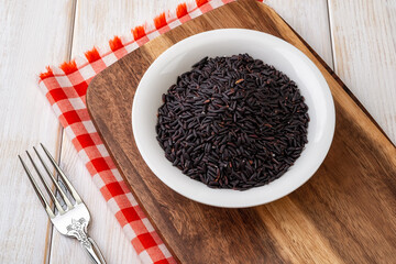 Raw dry black rice grains in a white ceramic bowl on a brawn wooden cutting board over wooden table. Unprocessed organic black rice as source of complex carbohydrates for dieting and healthy eating.