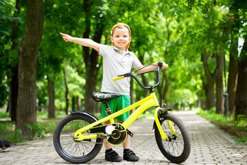A small smiling five-year-old boy in a yellow cap stands on the path with his bright lemon yellow bicycle and points to the side.