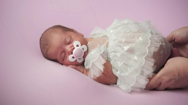 A little baby girl in a white dress lies on her stomach. Mom soothes the baby. A child with a pacifier in his mouth lies on a lilac background.