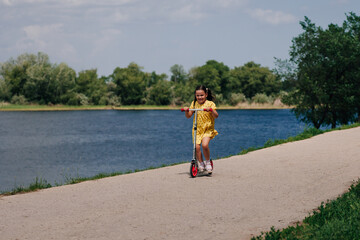 a girl in a yellow dress learns to ride a scooter on a narrow path with a river and an island, a summer blue sky on a sunny day. 