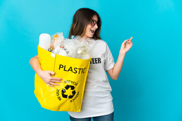 Woman holding a bag full of bottles to recycle isolated on blue background pointing finger to the side and presenting a product