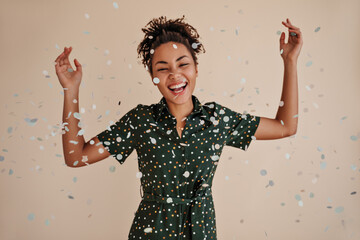 Joyful black young woman dancing under confetti. Studio shot of amazing laughing african american girl isolated on beige background.