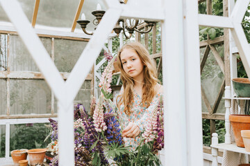 beautiful teen girl in a dress does summer bouquet of lupins in a white frame of veranda with flowers, a concept of summer vacation and a simple living