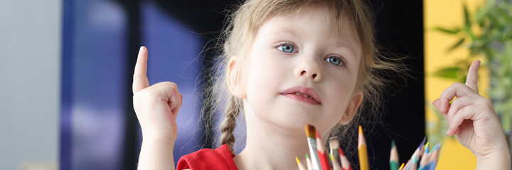Little girl sits at table with pencils and counts numbers on her fingers