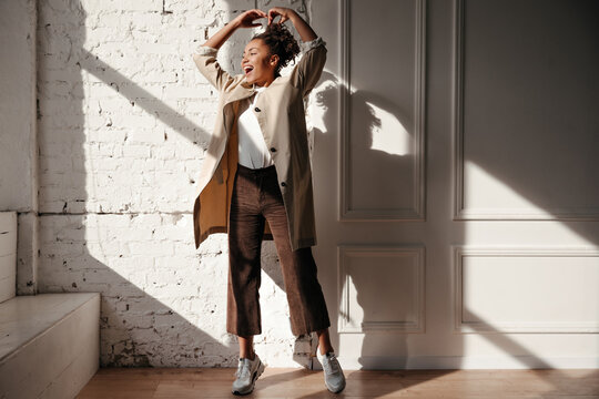Full length view of black lady in trench coat posing with hands up. Indoor shot of winsome african american girl enjoying sunlight.