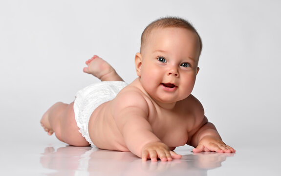 baby infant lies on his stomach in one diaper, lifted his bare feet up, happily looking at the camera. on a light background.