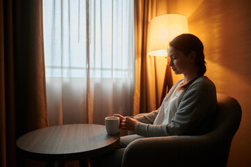 A young woman is sitting in an armchair in a cozy room with a cup of coffee. Beautiful interior. Curtains on the windows. There is a floor lamp in the corner of the room. Home comfort.