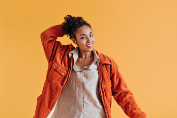 Stunning african american woman touching curly hair. Studio shot of gorgeous black girl wearing windbreaker on yellow background.