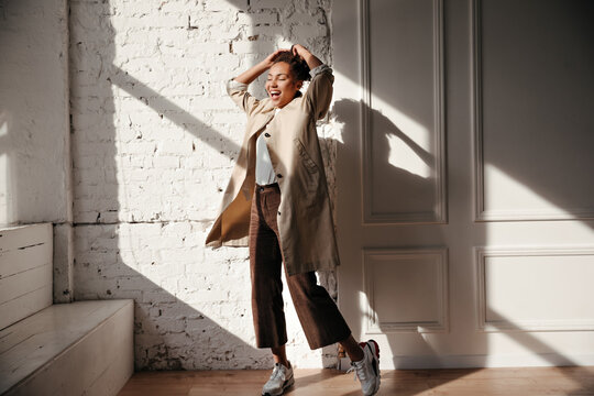 Full length view of ecstatic black woman in trench coat. Indoor shot of blissful african american girl enjoying sunshine.