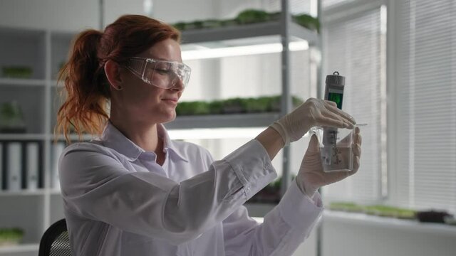 biological research genetic engineering laboratory, female laboratory worker in protective goggles conducts research with water to irrigate micro-greens and drips reagent into flask background of