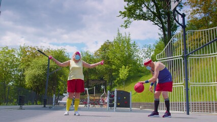 Active senior handsome man and woman playing basketball outdoors on sports playground court. Elderly old grandparents couple wearing medical mask during coronavirus pandemic. Recreational activity