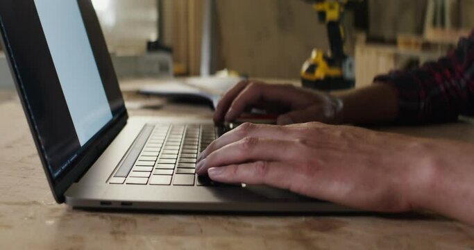 man with fair skin is typing on laptop, hands close-up, in a carpentry workshop