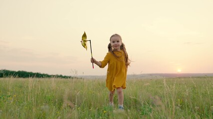 Little girl, daughter runs with toy windmill in her hand on summer field at sunset. Family vacation in nature. Happy child playing with toy turbine outdoors in spring park. Childhood, children