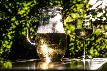Glass and jug of wine with water drops on them in sunlight on a wooden table, shooting from the side, close-up, against the background of foliage