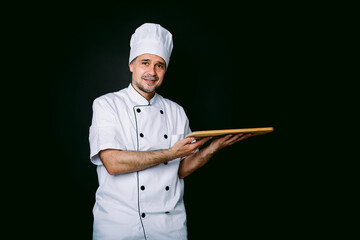 Chef cook wearing cooking jacket and hat, holding a wooden tray, on black background