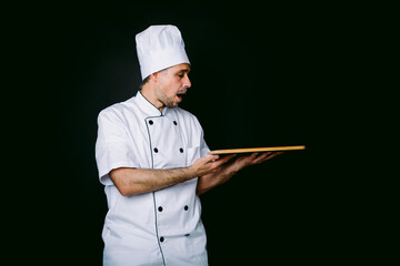Chef cook wearing cooking jacket and hat, surprised, holding wooden tray, on black background