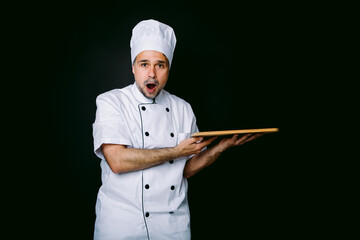 Chef cook wearing cooking jacket and hat, surprised, holding wooden tray, on black background