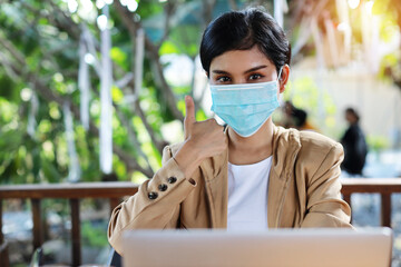 Fototapeta na wymiar Young asian woman hands in casual dress with protect mask for healthcare, sitting in coffee shop and using on smartphone and working on laptop computer and showing thumb up symbol