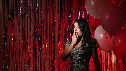 Holiday surprise. Amazed woman. Unexpected situation. Excited beautiful lady elegant dress looking shocked on red shimmering cascade curtain colorful balloons decor copy space.