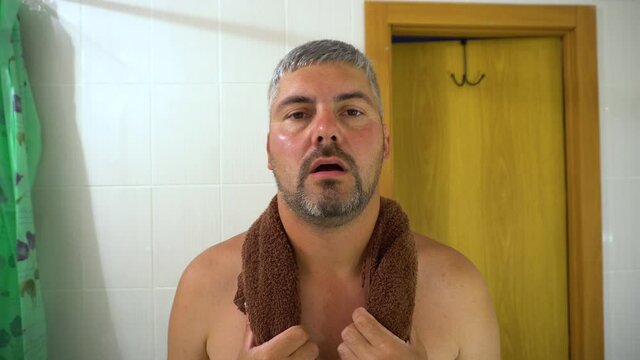 A grown man is standing in the bathroom. There is a towel around my neck. Tired look. After taking a shower. After workout. He has a beard on his face. White hair. Close-up.