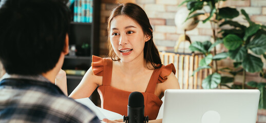 Cheerful young Asian female podcaster holding a questionnaire while interviewing male guest at workspace with mic and laptop