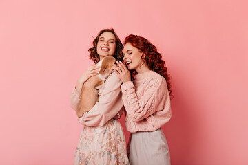 Studio shot of two friends with dog. Curly girls playing with puppy on pink background.