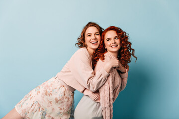 Laughing young woman embracing best friend. Beautiful girls having fun on blue background.