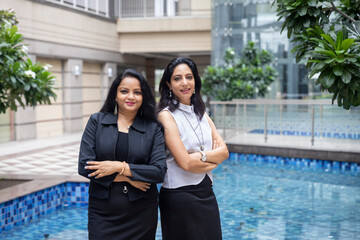 Two smart Indian woman standing side by side with their arms crossed in an urban corporate setting.