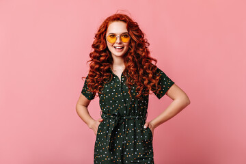Appealing ginger girl standing with hands on hips. Happy curly young woman in sunglasses posing on pink background.