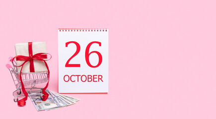 A gift box in a shopping trolley, dollars and a calendar with the date of 26 october on a pink background.