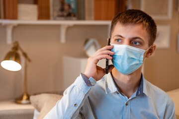 a young man of Caucasian appearance, in casual clothes, talking on the phone in a protective mask while sitting on the couch at home. The concept of remote communication