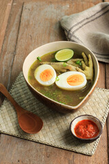 Indonesian Traditional Food Named Soto Ceker Ayam (Chicken Soup with Chicken Leg) on Brown Ceramic Bowl, with Sambal, Boiled Egg, and Lime. On Rustic  Wooden Table