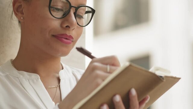 Tilt-up slowmo close-up of confident businesswoman in stylish eyeglasses taking notes in her planner in urban environment