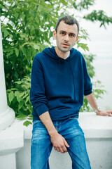 thin handsome young man in a blue hoodie and jeans is sitting against a background of greenery. portrait of a man of 30-40 years old. selective focus
