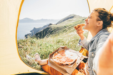 Active young woman brunette makes selfie while holding pizza slice in hand and sitting in yellow tent against rocky hill edge and misty landscape close view