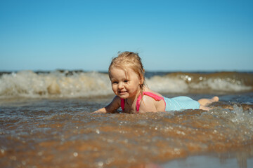 Fototapeta na wymiar Cute Little caucasian blond girl lying in sea in waves. Vacation with kids. Sunny day on the beach