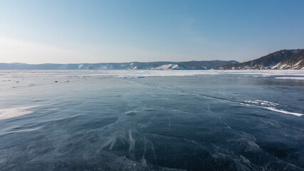 Cracks and some snow are visible on the smooth ice of the frozen lake. In the distance, a mountain range against a blue sky. Baikal