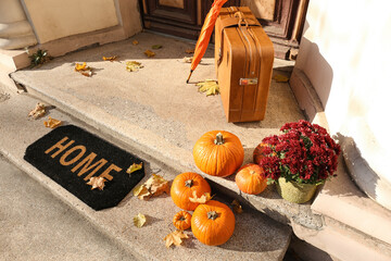 Doormat with umbrella, suitcase and pumpkins near entrance of house on autumn day