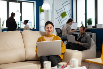 Businesswoman sitting on couch holding laptop, smiling at camera while diverse colleagues working in background. Multiethnic coworkers talking about start up financial company in modern business