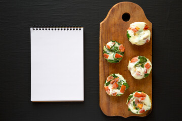 Homemade Egg White Breakfast Cups with Spinach and Tomato on a rustic wooden board, blank notepad on a black background, top view. Flat lay, overhead, from above.